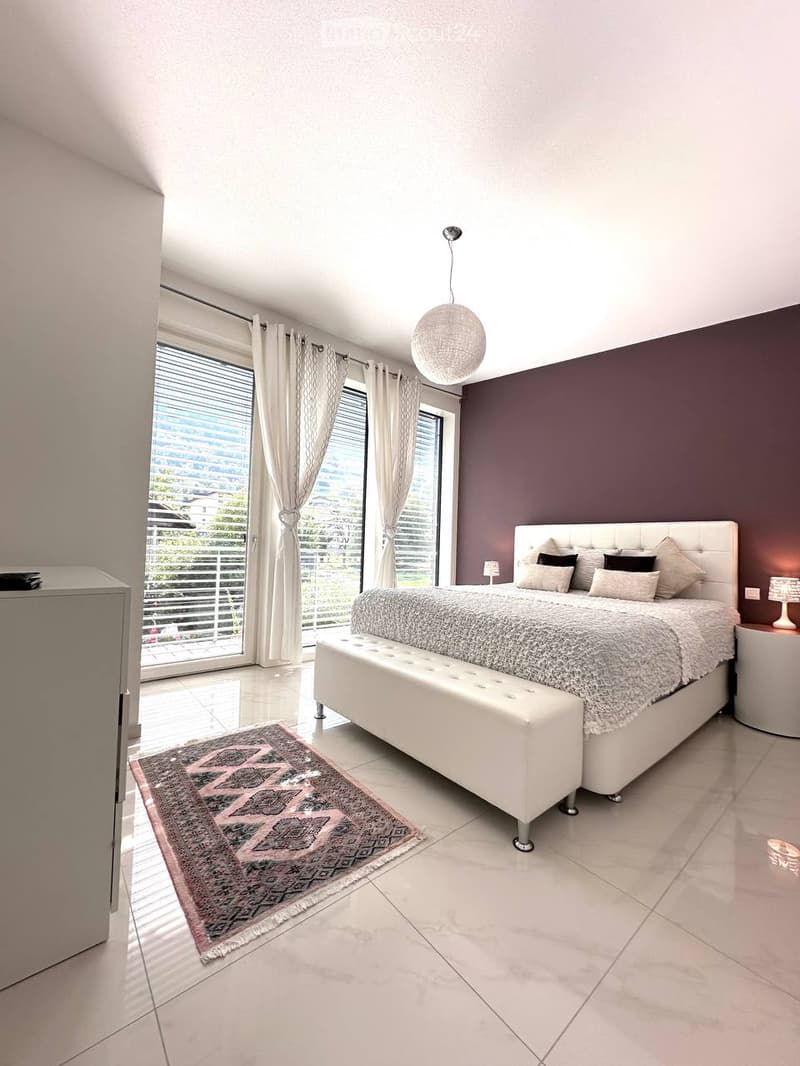 Modern bedroom with a large bed, bench at the foot, side table, and a patterned rug on the floor. Large windows with curtains offer a view of the outside.
