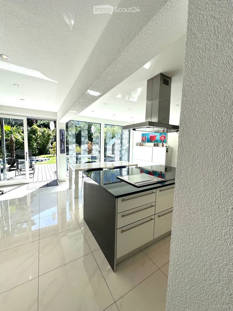 Modern kitchen with white cabinets, black island, stainless steel range hood, and a large island table. The space features large windows and a sleek, contemporary design.