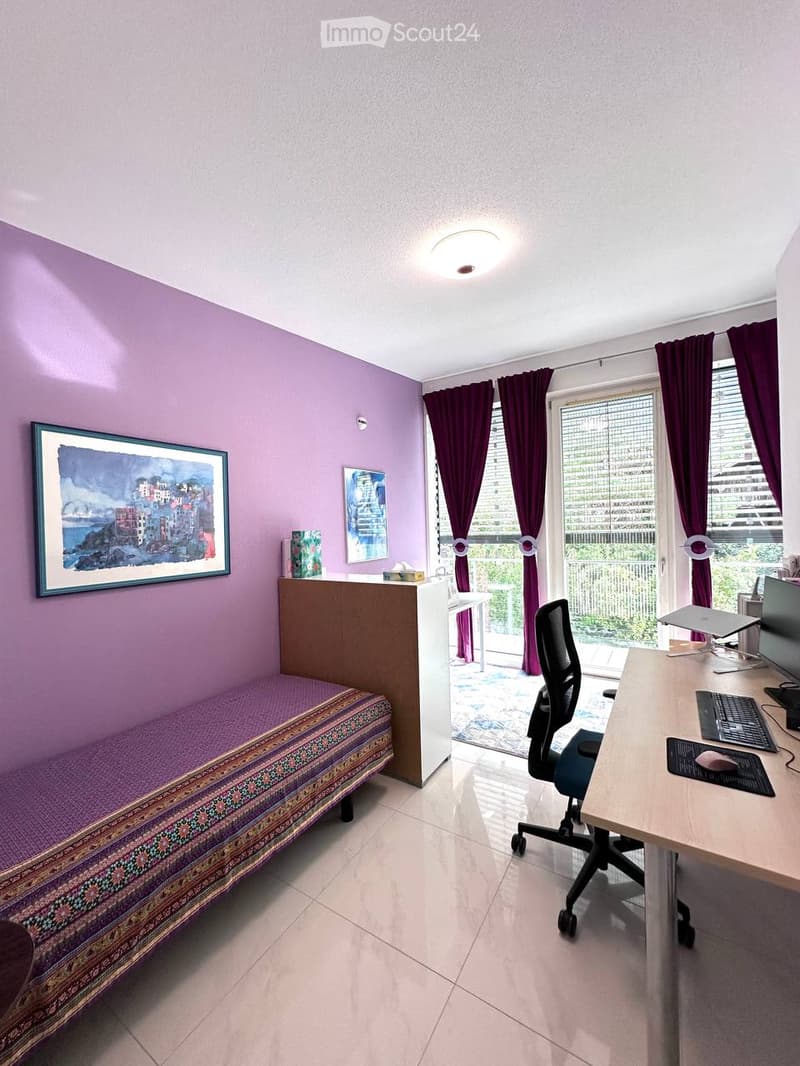 A well-lit room with a purple accent wall, a sofa, a desk with a computer setup, and a large window with purple curtains leading to a balcony.