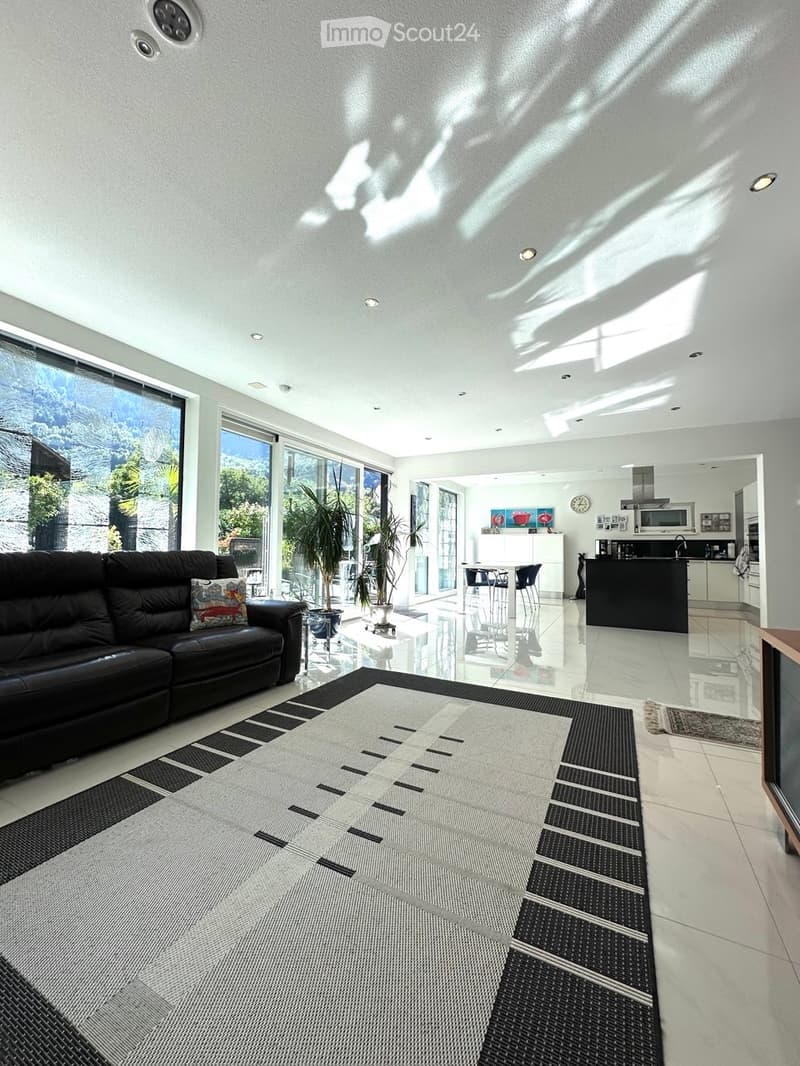 Modern living room with a black leather sofa, coffee table, potted plants, and a dining area with a table and bar stools. The kitchen features a black island with a microwave and a clock on the wall.