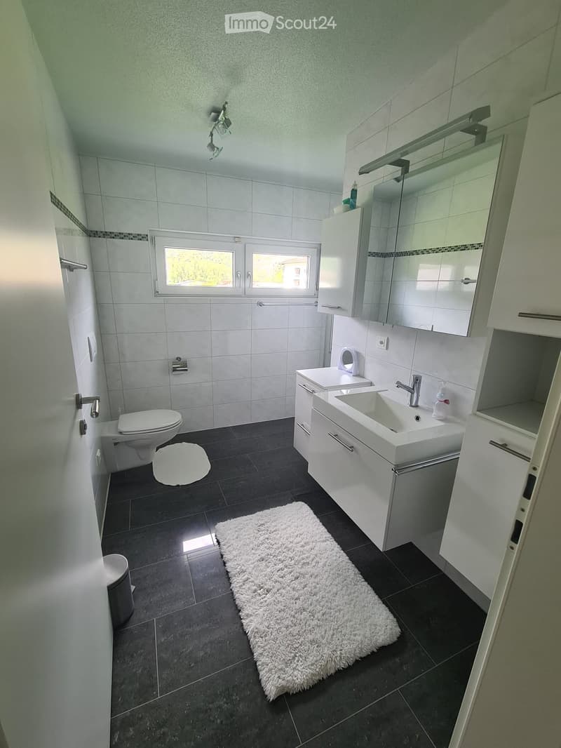 Modern bathroom with white tiles, dark floor, double sink, toilet, mirror, and a fluffy white rug.
