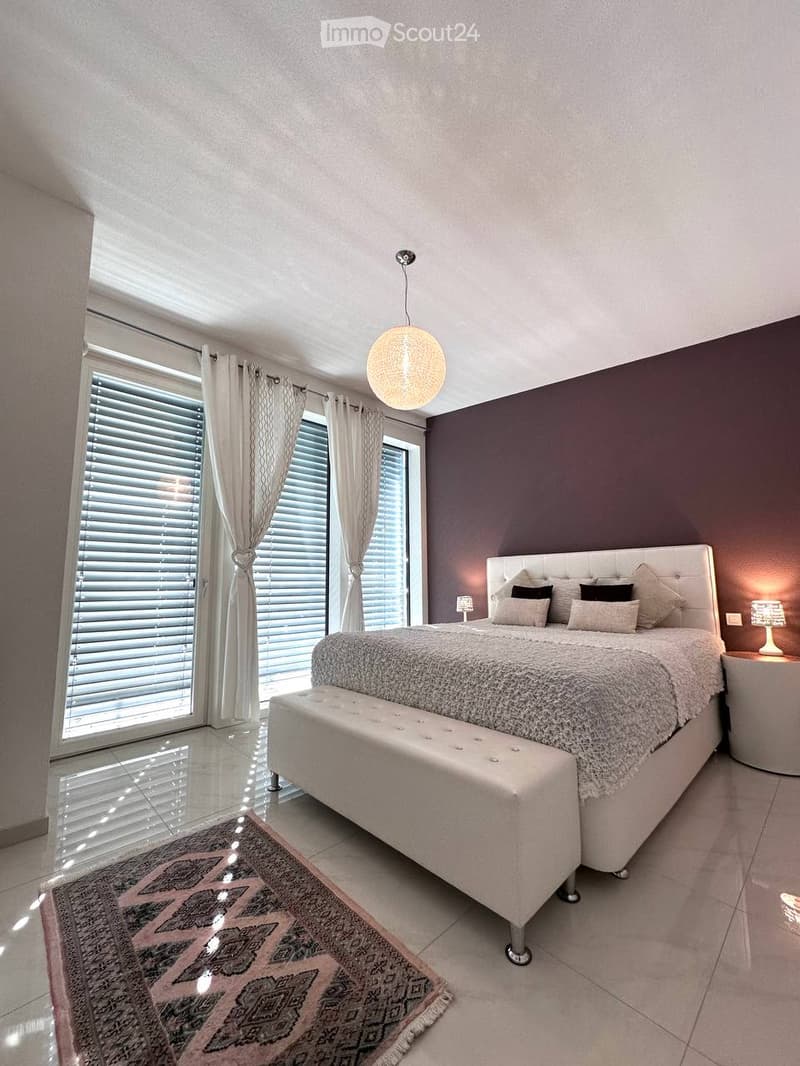 Modern bedroom with a large bed, bench at the foot, nightstand, and large windows with curtains.