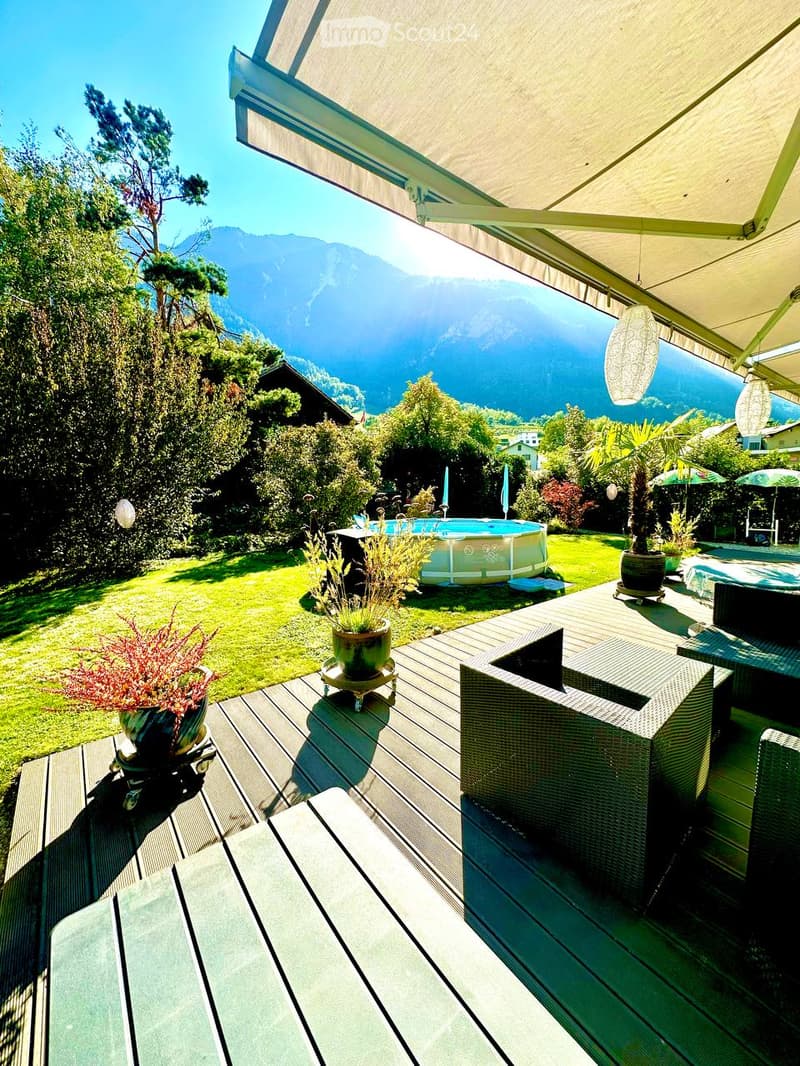 Outdoor patio with wooden decking, wicker chairs, a table, potted plants, and a swimming pool, overlooking a scenic mountain view.