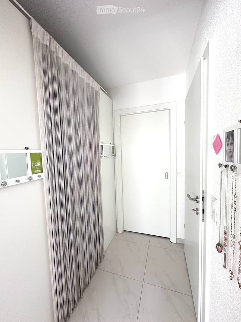 A clean, modern hallway with white walls, tiled flooring, and a closed door at the end. A curtain is on the left side, and a small shelf with a mirror and decorative items is visible.