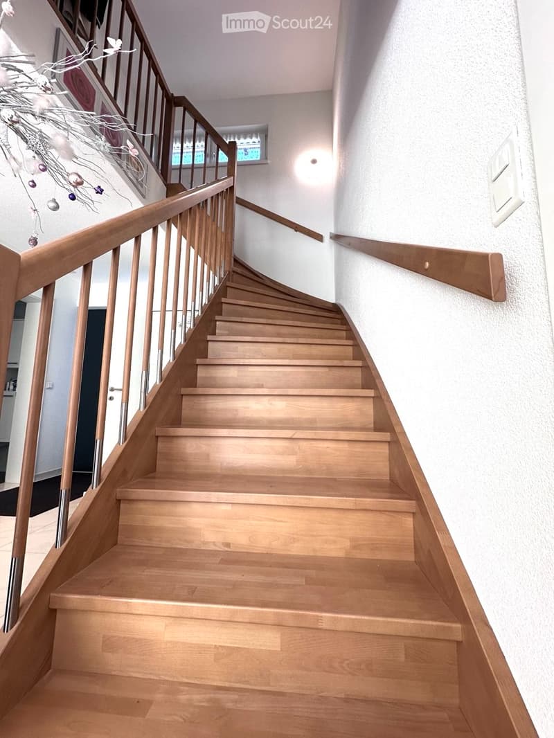 Wooden staircase with a wooden handrail and metal balusters, leading upwards. The walls are white, and there is a small decorative branch on the left side.