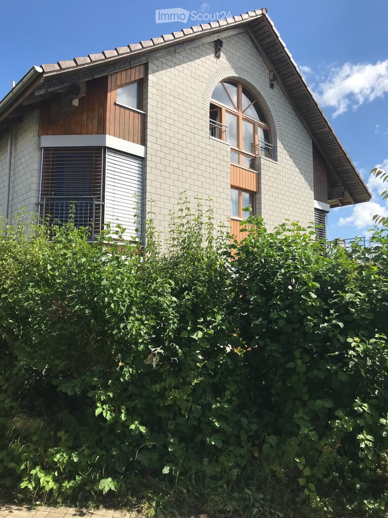 Two-story house with white brick facade, wooden accents, and multiple windows, partially obscured by dense green foliage.
