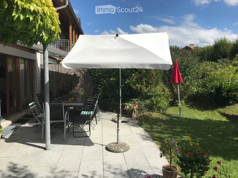 Outdoor patio area with a large umbrella, a table, several chairs, and surrounding greenery.