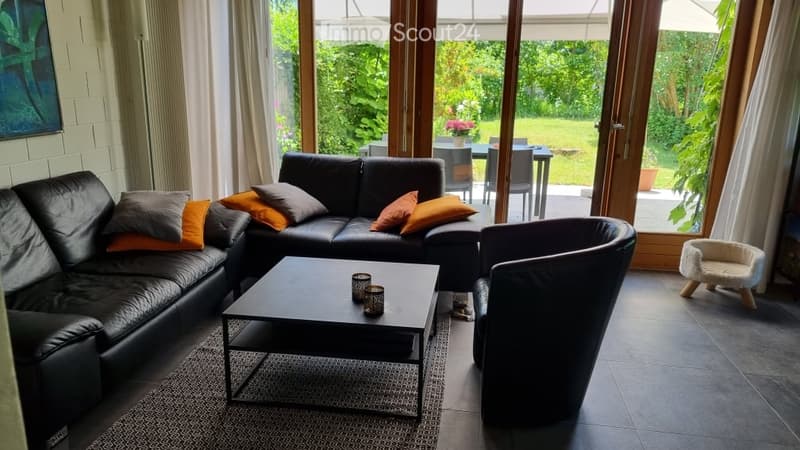 Modern living room with large windows offering a view of a garden, featuring a black leather sofa with colorful cushions, a coffee table, an armchair, and a small pet bed in the corner.