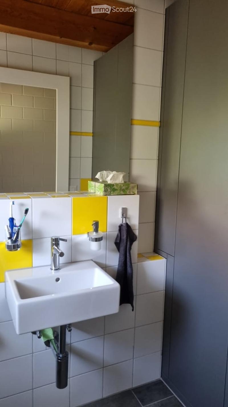 Modern bathroom with a white sink, chrome faucet, mirror, toothbrush holder, and a towel hanging on a rack. The walls are tiled with a yellow accent border.