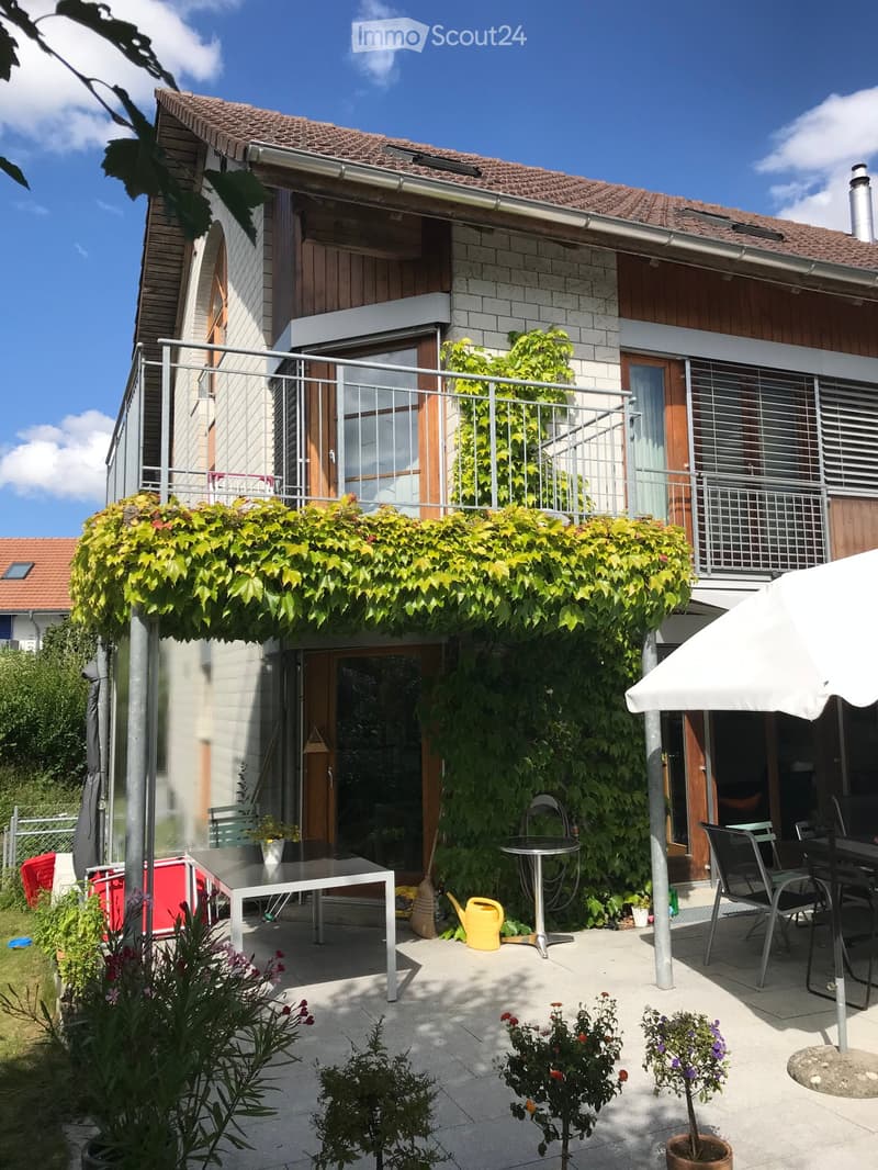 Balcony area with a table, chairs, umbrella, potted plants, and a watering can. The balcony is attached to a house with a wooden facade and a tiled roof.
