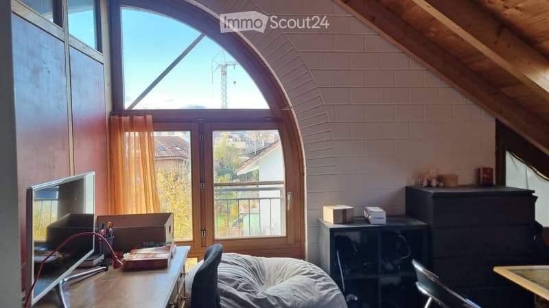 Bright attic room with slanted wooden ceiling, large arched window, and balcony access. Contains a screen, chair, table, and small cabinet.