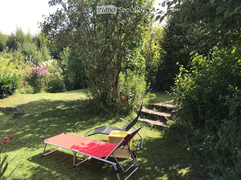 A serene garden with a sun lounger, a chair, and lush greenery, including trees and flowering plants.
