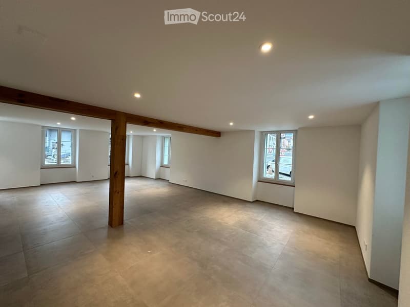 Empty room with tiled floor, white walls, large windows, and exposed wooden beams.