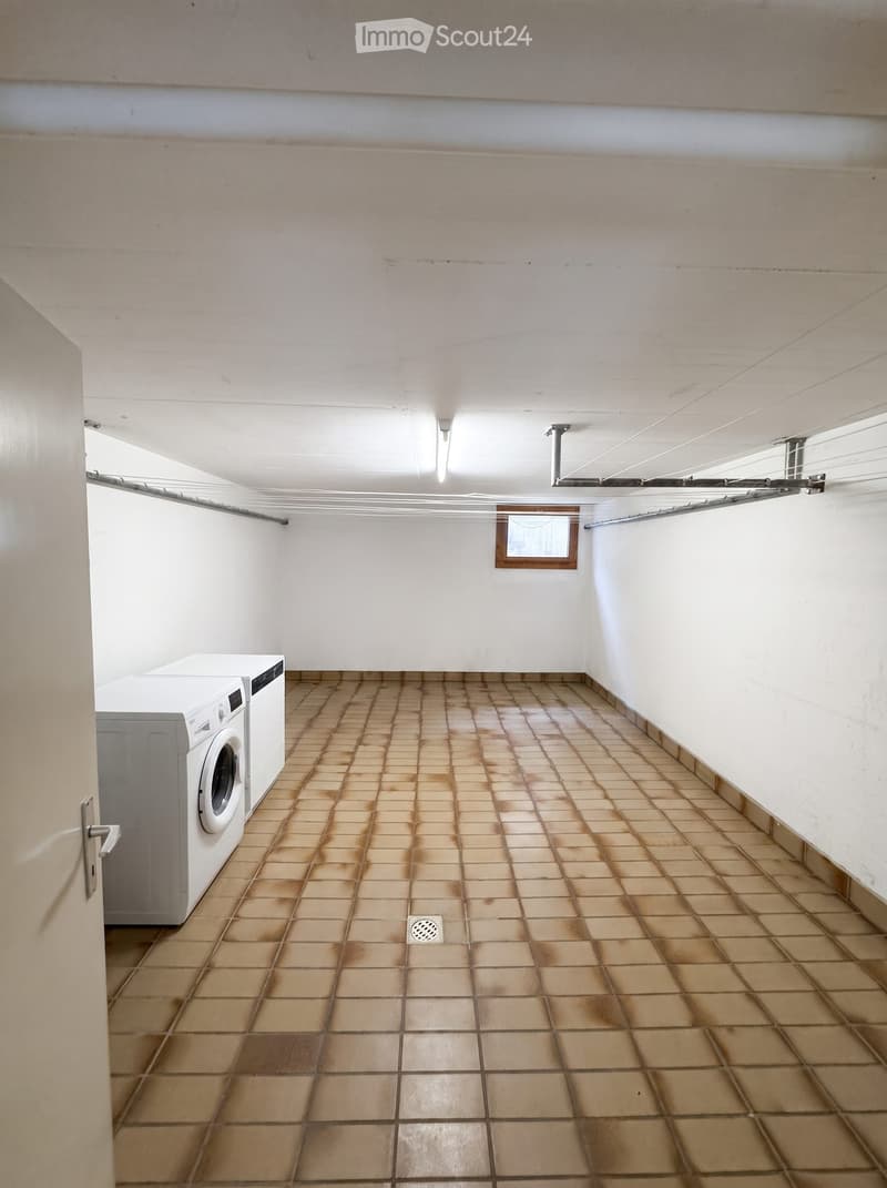 Laundry room with tiled floor, white walls, and a washing machine. Features a small window and a drain in the center of the floor.