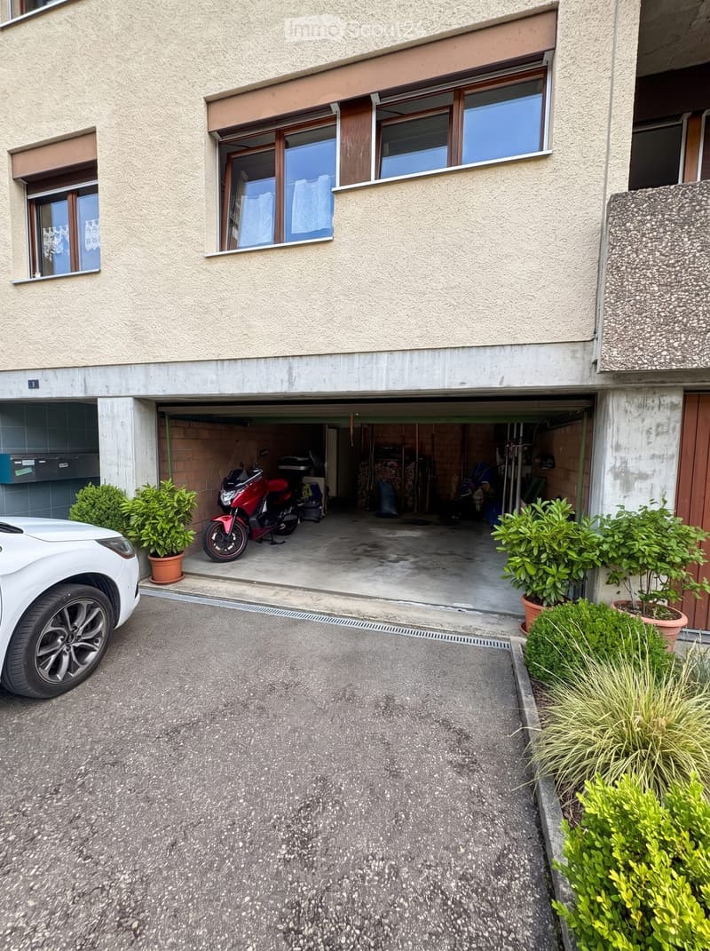 Two-story building with a garage, white car parked outside, and potted plants near the entrance.