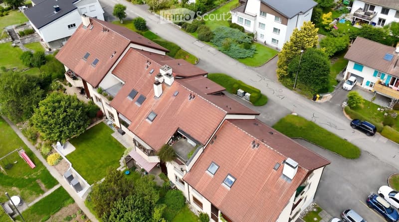 Aerial view of a residential area with multiple houses, red-tiled roofs, green lawns, and parked cars.