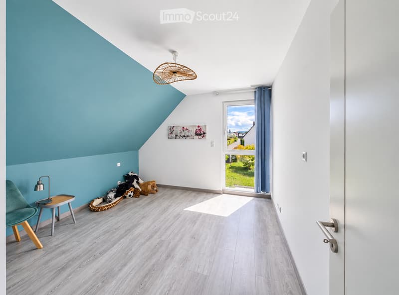 Bright, modern room with slanted ceiling, wooden flooring, large window with view, and colorful decor including a green chair, small table, lamp, and toys.