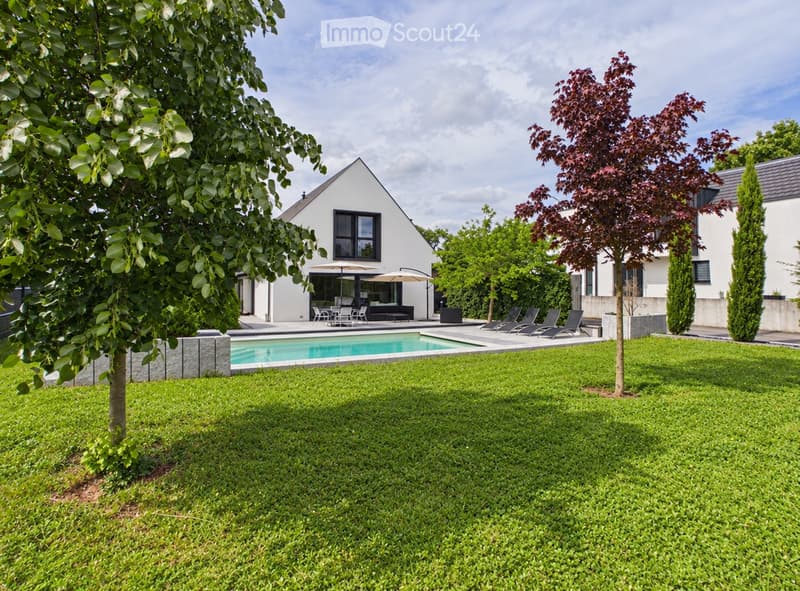 Well-maintained garden with a swimming pool, lounge chairs, and outdoor dining area under umbrellas, surrounded by trees and a modern white house.