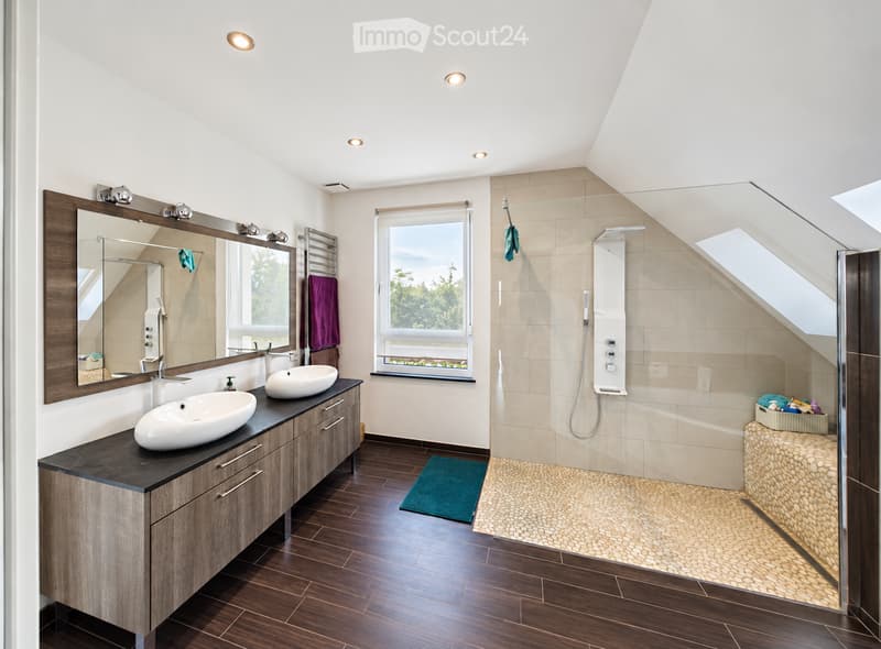 Modern bathroom with double sinks, large mirror, wooden vanity, and a shower area with a glass enclosure. The space features a skylight window and a view of greenery outside.