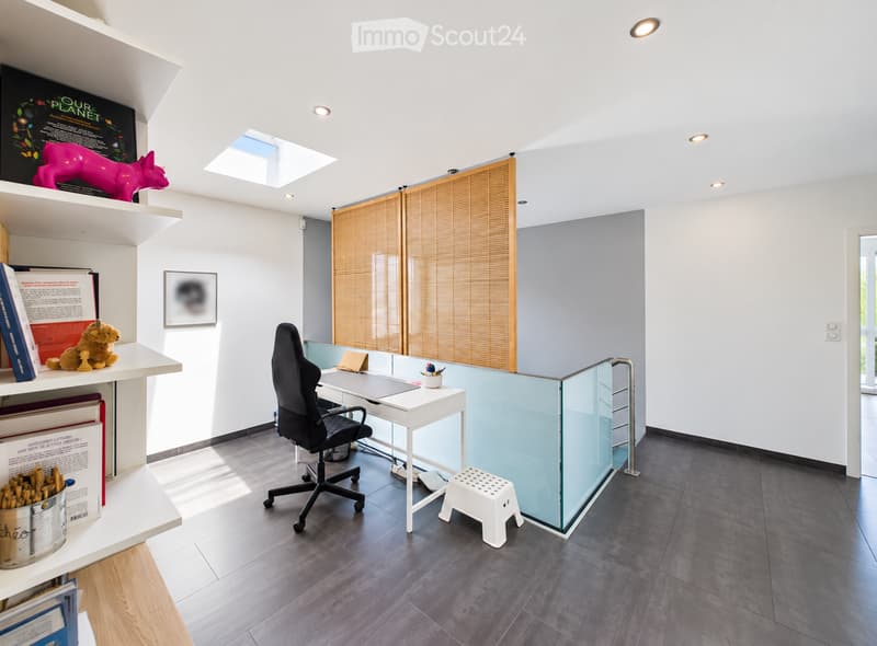 Modern office space with a white desk, black office chair, glass partition, and shelves holding books and decorative items.
