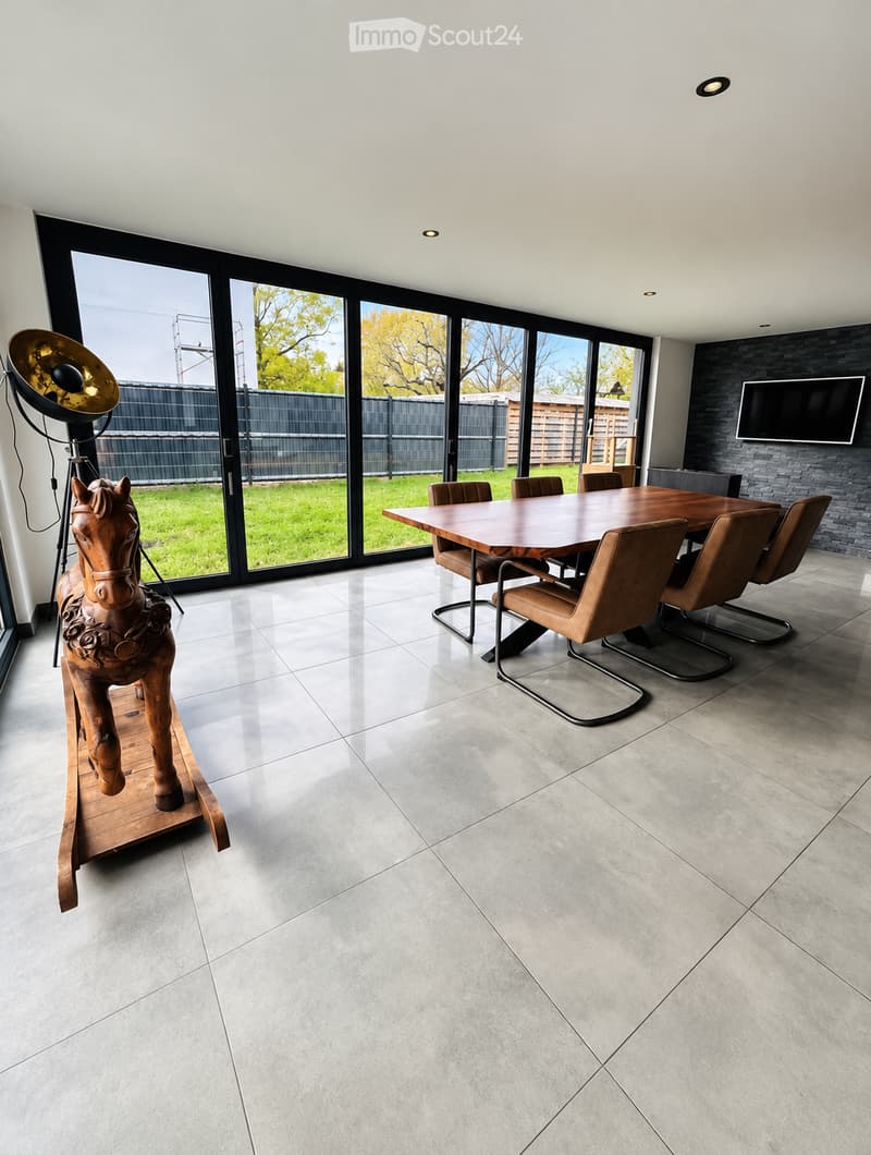 Modern dining area with a large wooden table, brown leather chairs, a decorative horse sculpture, and a wall-mounted screen. Large windows offer a view of a green lawn and outdoor structures.
