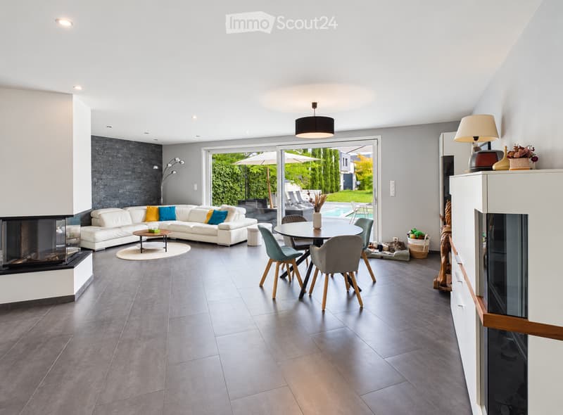 Modern living room with a white sectional sofa, fireplace, dining area, and large windows offering a view of the garden and pool.