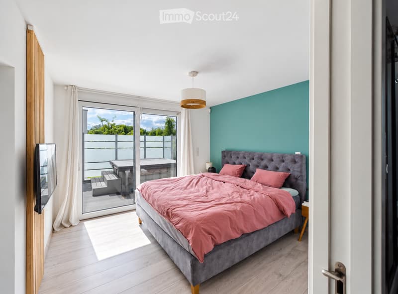 Modern bedroom with a grey upholstered bed, pink bedding, a nightstand, a wall-mounted mirror, and a large window with a view to a balcony area.