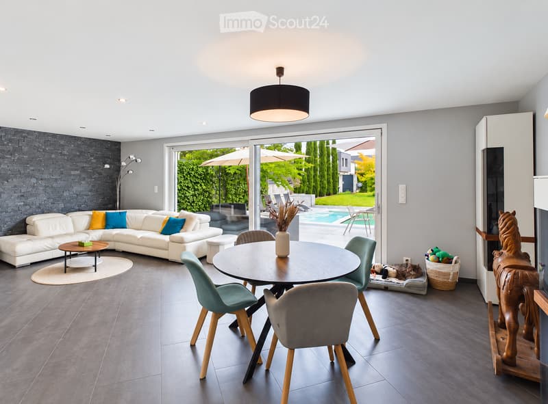 Modern living and dining area with a white sectional sofa, colorful cushions, a round coffee table, a dining table with chairs, a cat bed, a decorative toy horse, and a view of a swimming pool through large glass doors.