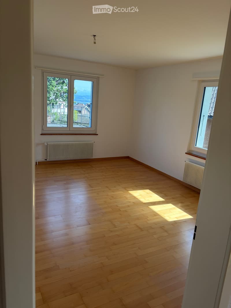 Empty room with wooden flooring, white walls, and large windows allowing natural light to enter. Radiators are visible along the walls.