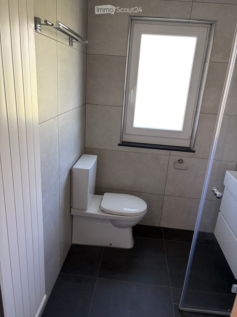 Modern bathroom with a toilet, sink, and a window above the toilet. The walls are tiled in a light gray color, and the floor is dark gray tiles.