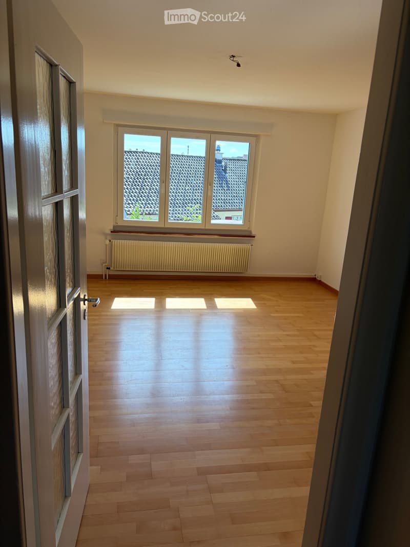 Empty room with wooden flooring, large windows overlooking a roof, radiator, and a door with glass panels.