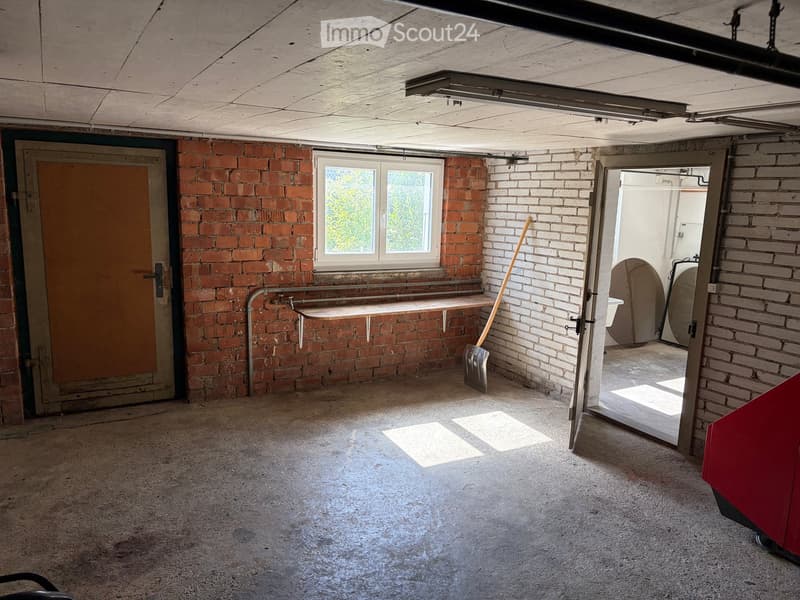 Empty room with exposed brick walls, concrete floor, single window, and a small shelf with a broom and shovel. No furniture or equipment present.