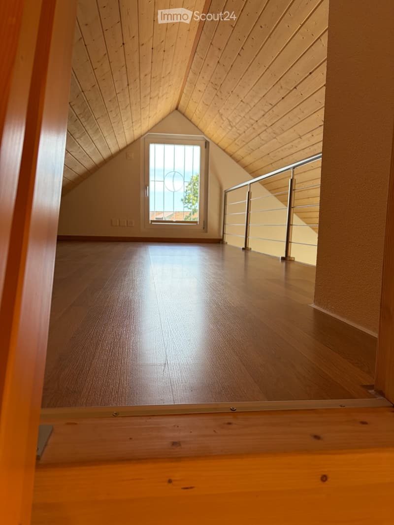 Empty attic space with wooden floors, slanted wooden ceiling, and a window with decorative glass. Metal railing visible on one side.