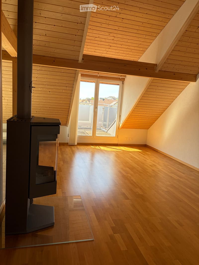 Empty attic room with wooden floors and slanted wooden ceilings, featuring a large window and a modern wood-burning stove.