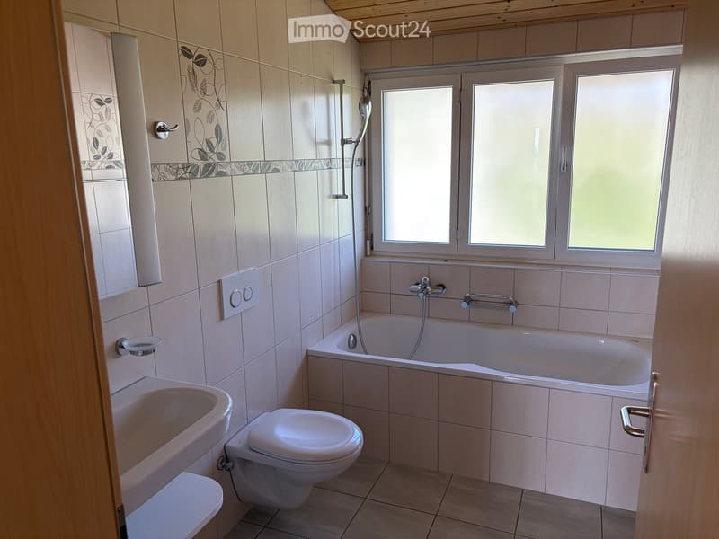 Modern bathroom with a bathtub, toilet, sink, and showerhead. Features tiled walls, a window, and a wooden door frame.