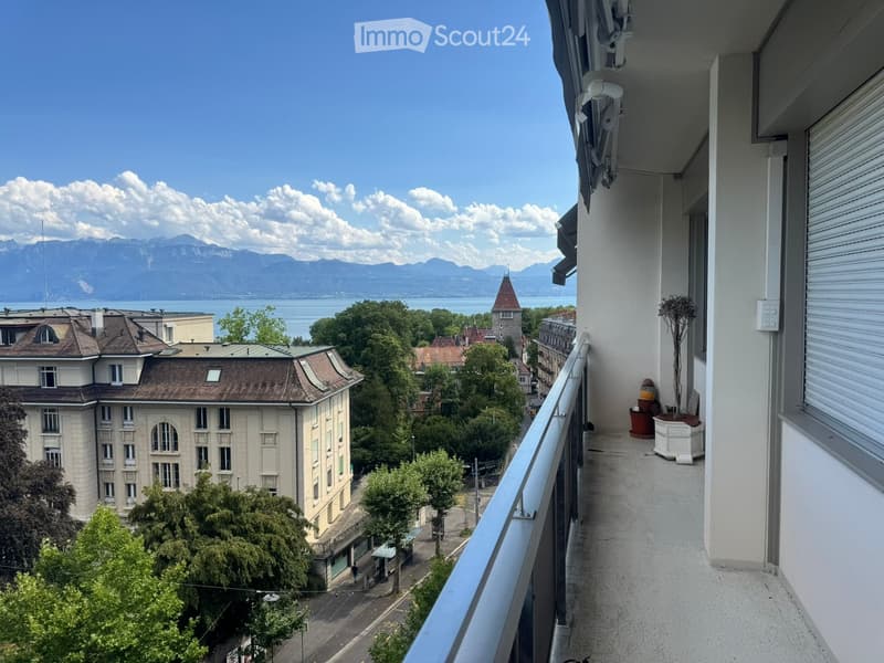 Balcony with a metal railing, overlooking a cityscape with buildings, trees, and a distant lake and mountains.