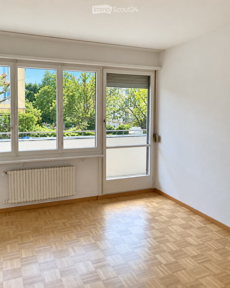 Empty room with wooden parquet flooring, white walls, large windows with blinds, radiator, and a view of greenery outside.