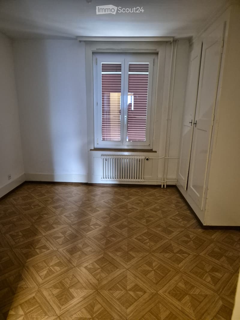 Empty room with wooden herringbone flooring, white walls, a window with blinds, and a radiator below it.