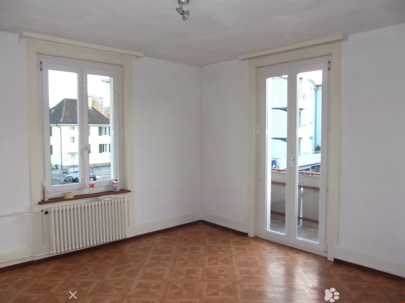 Empty room with wooden flooring, white walls, large window, and sliding glass door leading to a balcony.