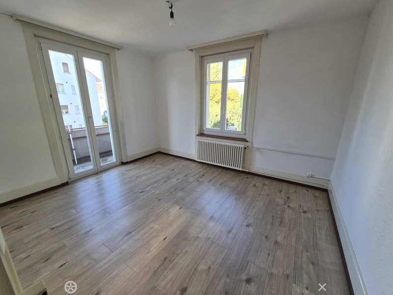 Empty room with wooden flooring, white walls, large window, and sliding glass door leading to a balcony.