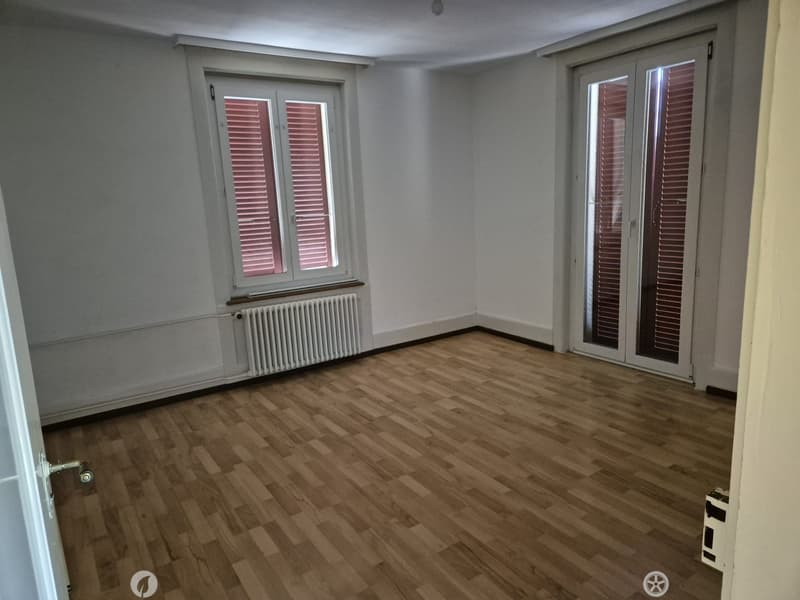 Empty room with wooden flooring, white walls, and large windows with blinds. Features a radiator and a door with a handle.