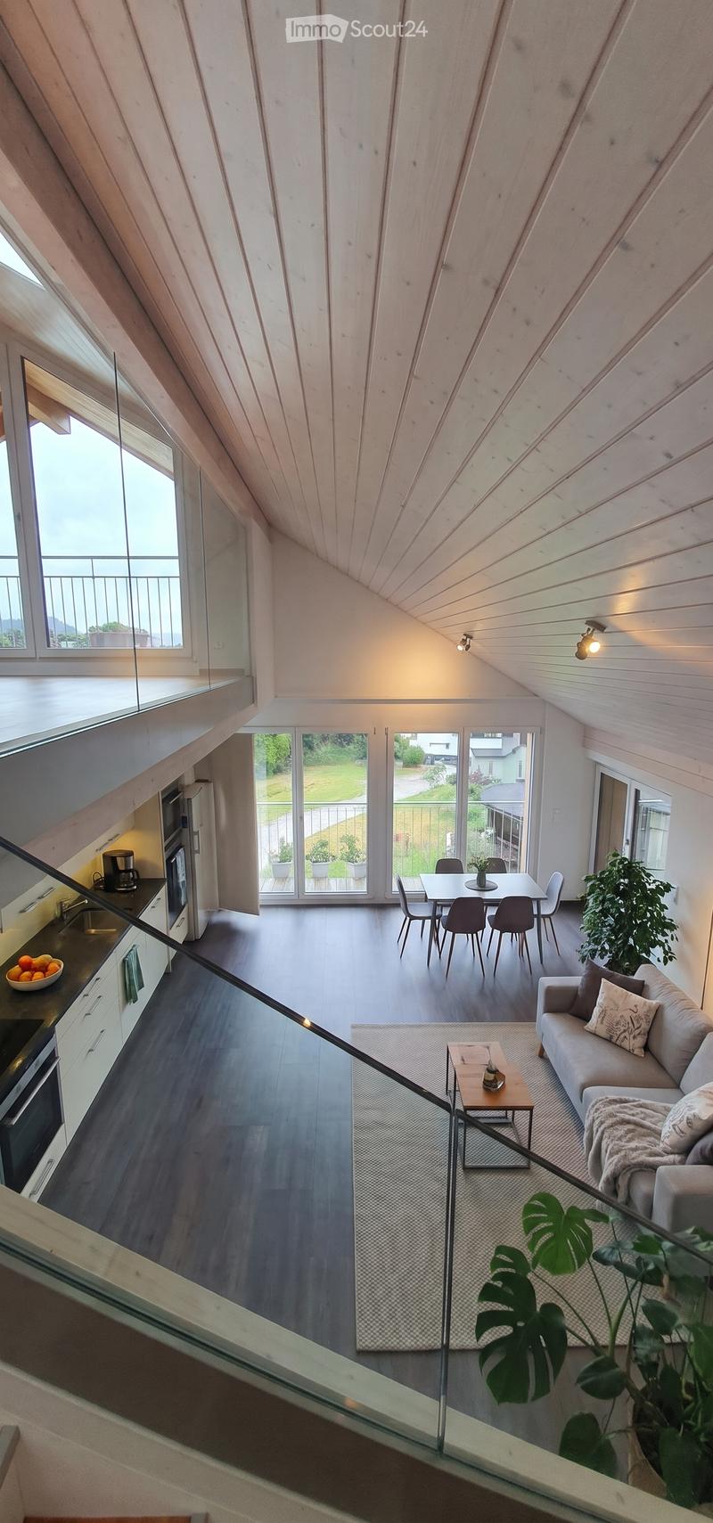 Open-plan living and dining area with a modern kitchen, sofa, coffee table, dining table, and chairs. Large windows offer a view of the garden.