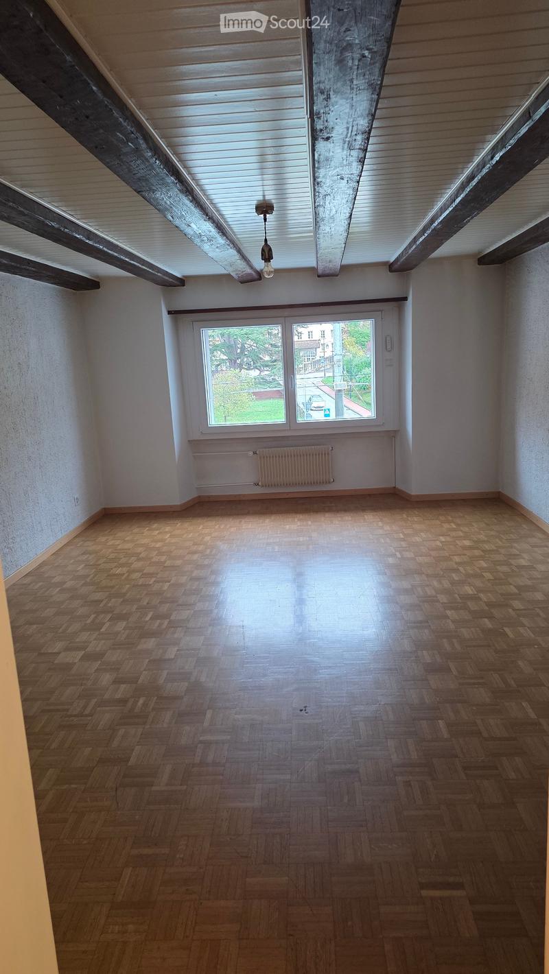 Empty room with wooden herringbone flooring, white walls, exposed wooden beams on the ceiling, and a large window with a view of the street.