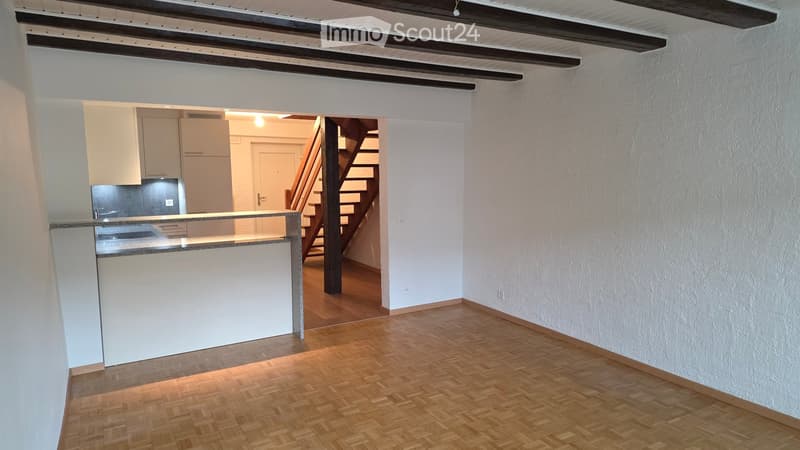 Empty room with wooden flooring, white walls, and a staircase leading to another area. There is a kitchen visible in the background with white cabinets and a countertop.