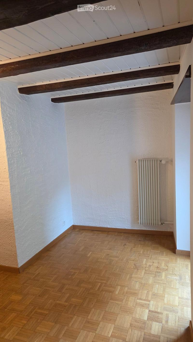 Empty room with wooden herringbone flooring, white walls, exposed wooden beams on the ceiling, and a radiator on one wall.