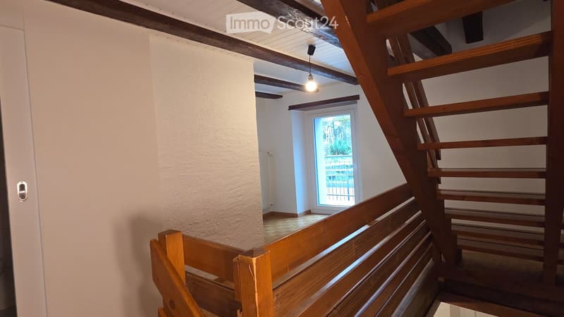 Wooden staircase with a wooden railing, white walls, and a hanging light fixture. A window at the end of the hallway provides natural light.