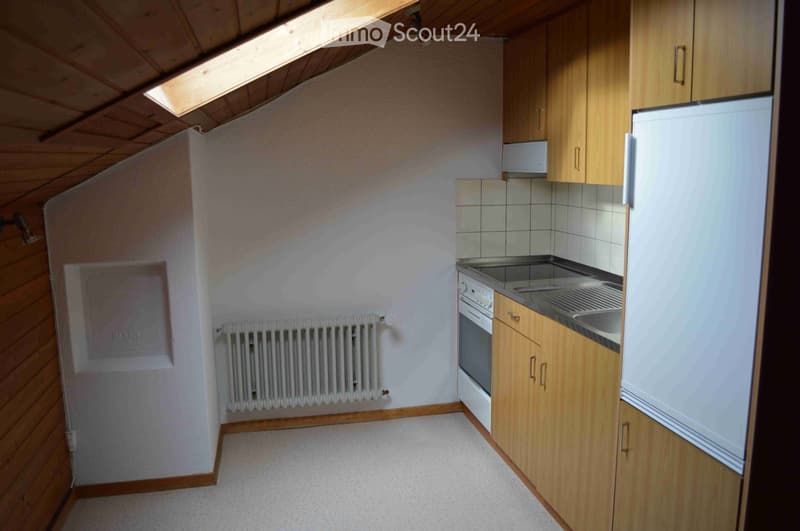 Compact kitchen with wooden cabinets, stainless steel sink, oven, and refrigerator. Light-colored walls and carpeted floor.