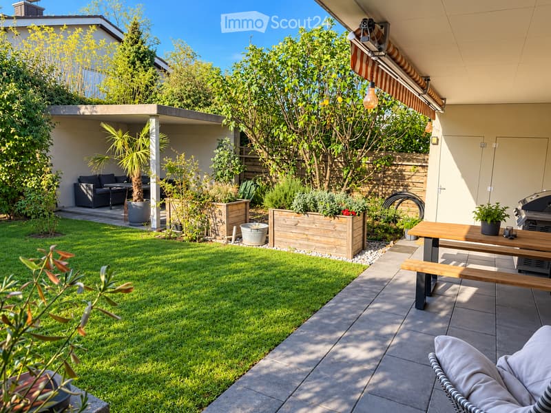 Well-maintained garden with a neatly trimmed lawn, wooden planters, a patio area with a wooden table and benches, and a covered seating area with a sofa and small table.