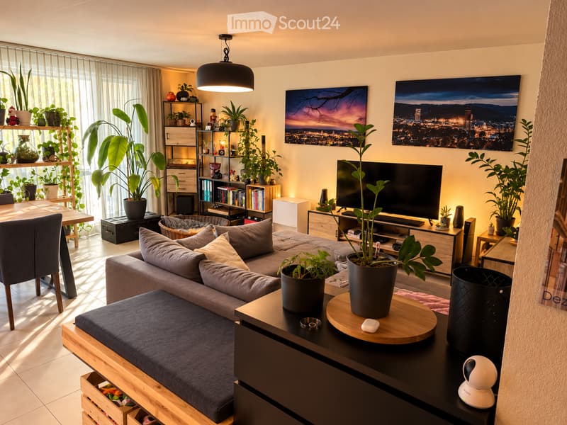 Modern living room with a grey sectional sofa, coffee table, dining area, shelves with plants and books, and large windows with sheer curtains.