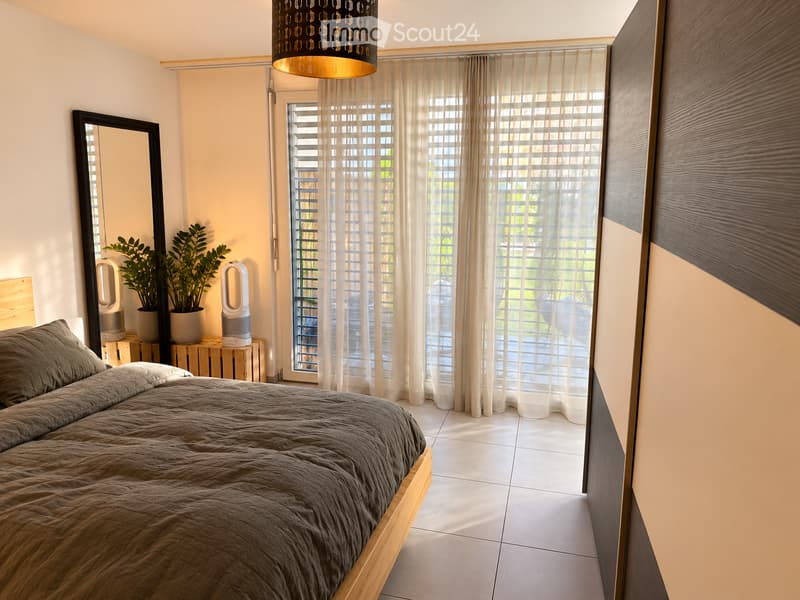 Modern bedroom with a wooden bed, grey bedding, large mirror, potted plants, speakers, and sheer curtains covering large windows.